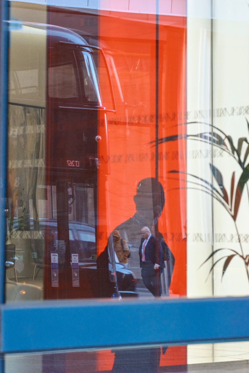 Abstract street reflection showing a man in a suit layered over a bright orange bus and architectural glass.