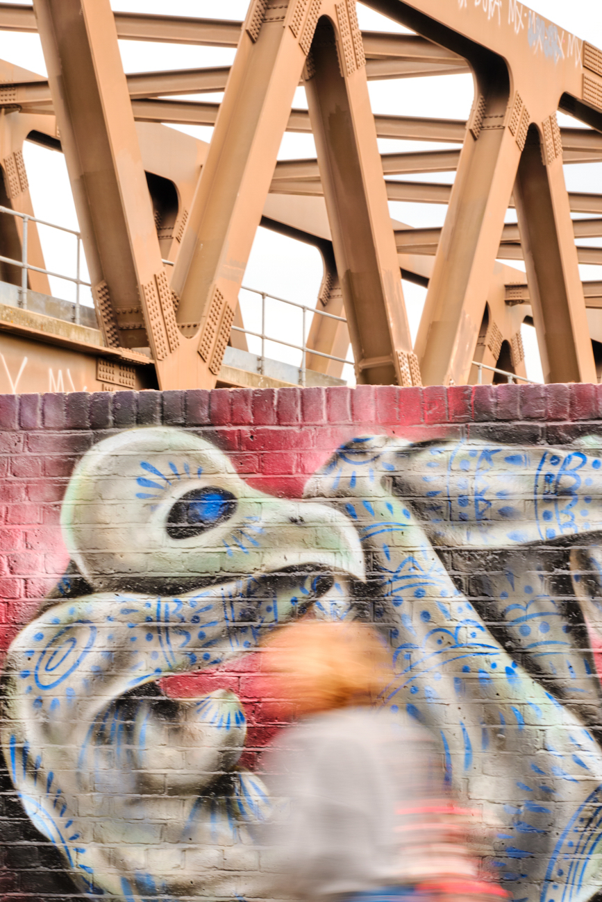 Motion blur of a person passing a vibrant graffiti mural of a bird beneath a heavy industrial steel bridge.