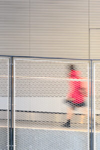 Blurred figure in red moving behind a white mesh fence against a ribbed neutral architectural background.
