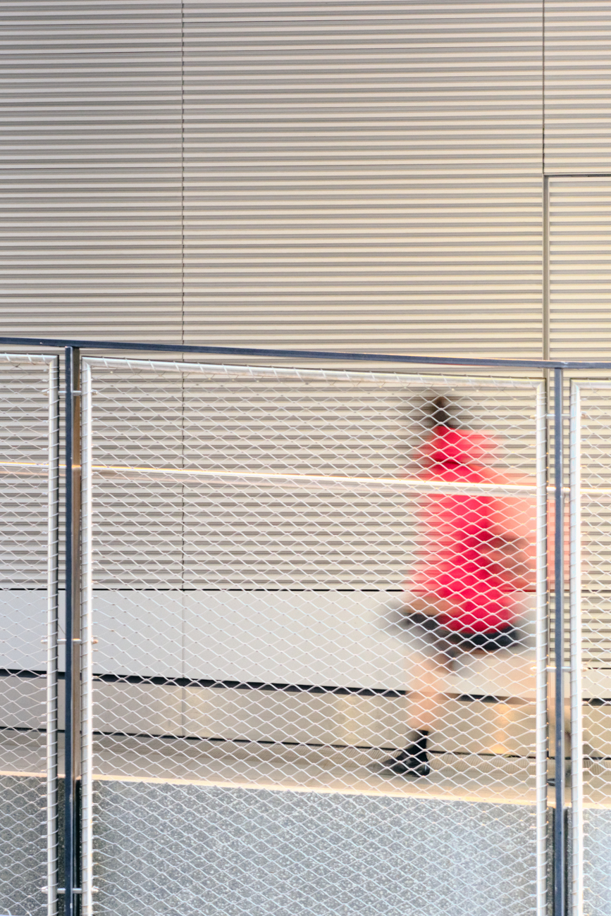 Blurred figure in red moving behind a white mesh fence against a ribbed neutral architectural background.