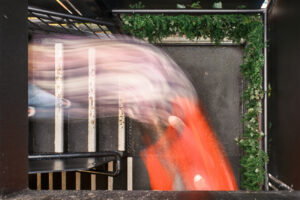 High-angle long exposure showing a ghostly red and white motion trail moving down industrial stairs with green foliage.