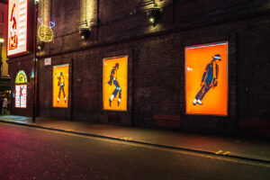 Night photography of an urban brick wall featuring three illuminated orange posters of a silhouetted figure.