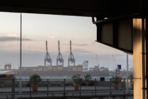 Industrial landscape of shipping cranes and container ships framed by a dark architectural structure.