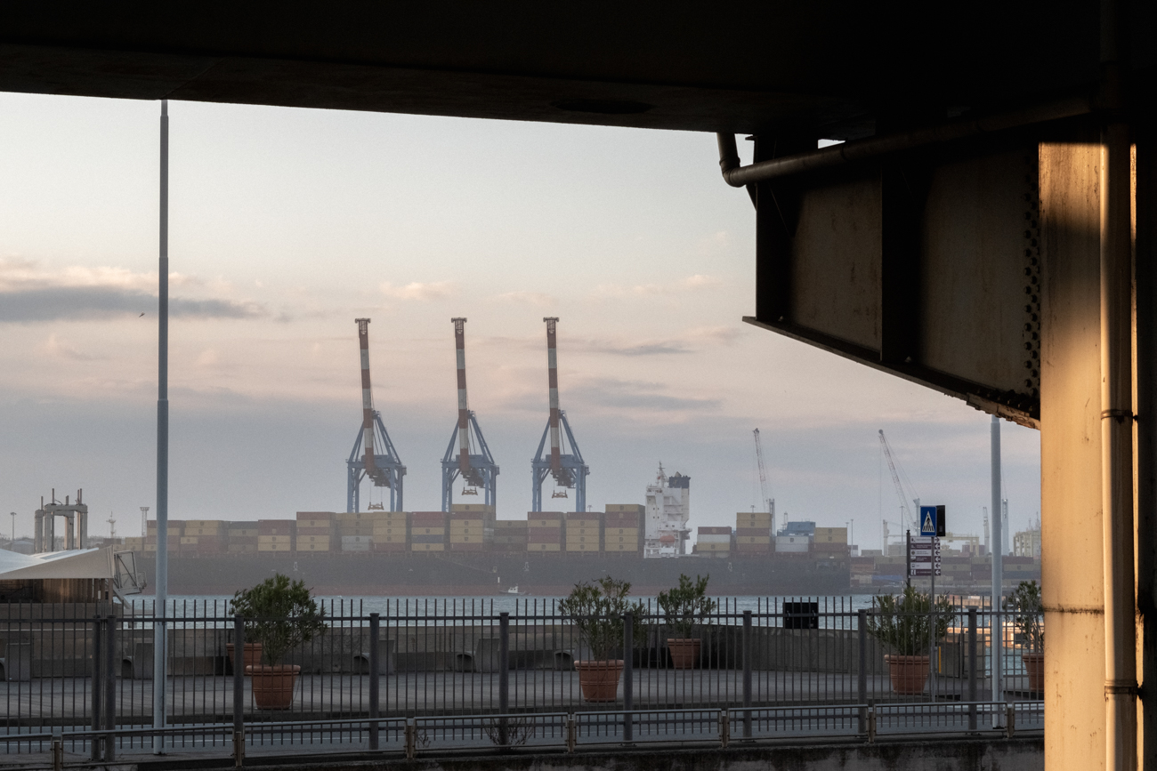 Industrial landscape of shipping cranes and container ships framed by a dark architectural structure.