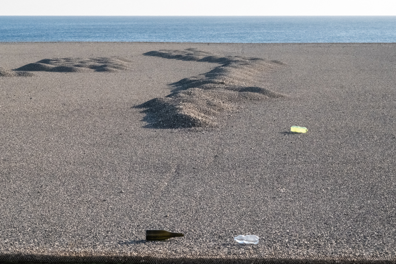 Fine art landscape of a pebble beach with discarded glass and plastic bottles, exploring human impact.