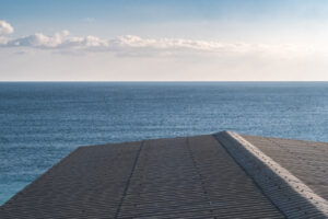Minimalist horizon study of a corrugated metal roofline meeting the sea under a clear sky.