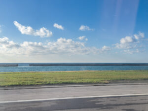 Coastal landscape with a green field, asphalt road, and breakwater wall stretching into the blue sea.